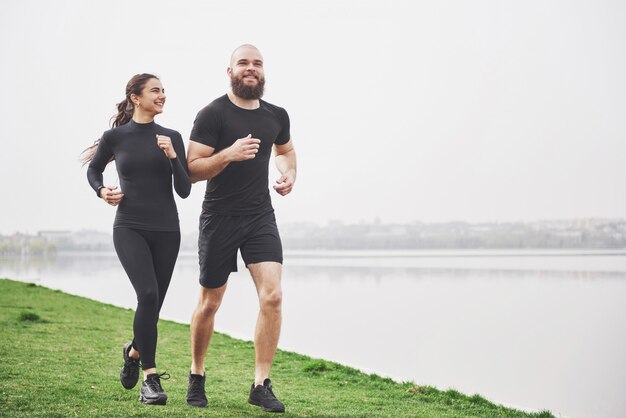 Couple jogging and running outdoors in park near the water. Young bearded man and woman exercising together in morning