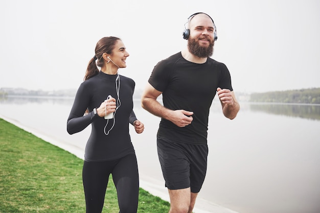 ジョギングや屋外の水の近くの公園で走っているカップル。若いあごひげを生やした男性と女性が朝一緒に運動
