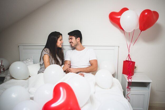 Пара смеется, лежа на кровати с воздушными шарами вокруг них