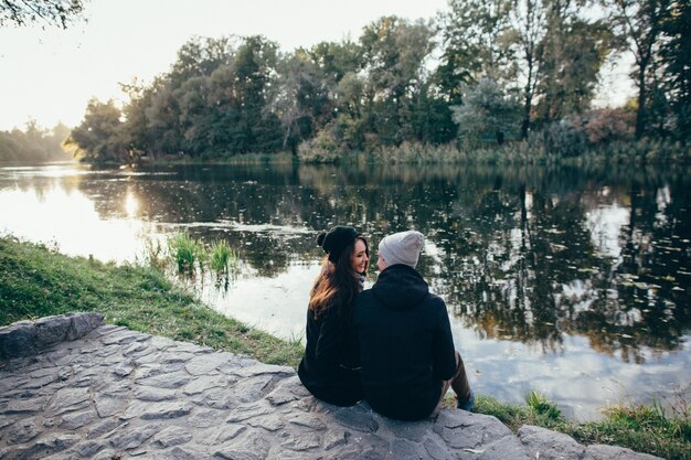 Влюбленная пара. Красивая пара в любви в теплой одежде сидит на берегу реки на закате.