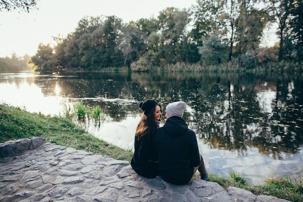 愛の恋人。暖かい服を愛する美しいカップルは、夕方に川沿いに座っています。