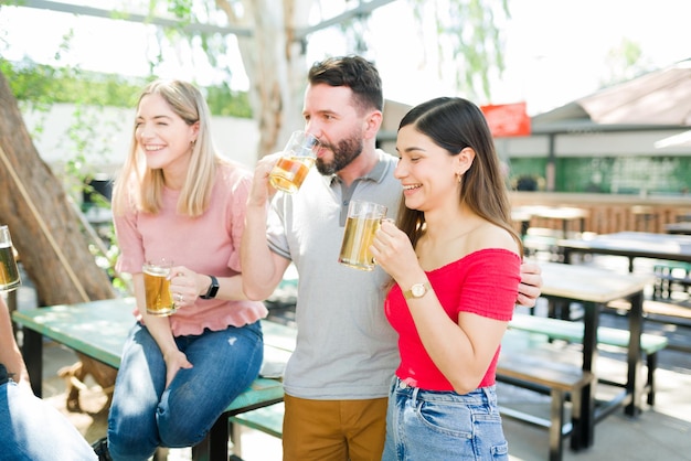 Couple in love hugging and laughing while drinking beer with some friends at an outdoor bar. Boyfriend and girlfriend hanging out with a group of friends