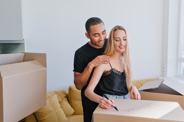 引っ越し用の箱に物を詰める愛のカップル、箱にラベルを貼る、妻と夫が新しい家を買って新しい場所に引っ越す。黄色のソファーと白い壁の部屋。