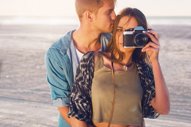 夕方のビーチ、流行に敏感な若い女の子と彼女のハンサムなボーイフレンドがレトロなフィルムカメラで写真を撮るポーズの愛のカップル。夕日の暖かい光。