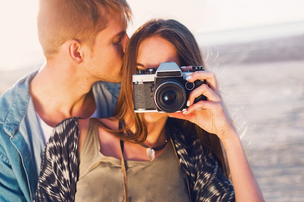 夕方のビーチ、流行に敏感な若い女の子と彼女のハンサムなボーイフレンドがレトロなフィルムカメラで写真を撮るポーズの愛のカップル。夕日の暖かい光。