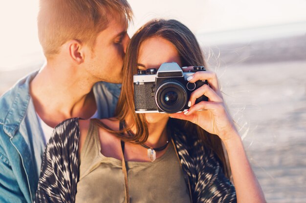 Пара в любви позирует на пляже вечером, молодая девушка битник и ее красивый парень фотографировать с ретро пленочной камеры. Закат теплым светом.