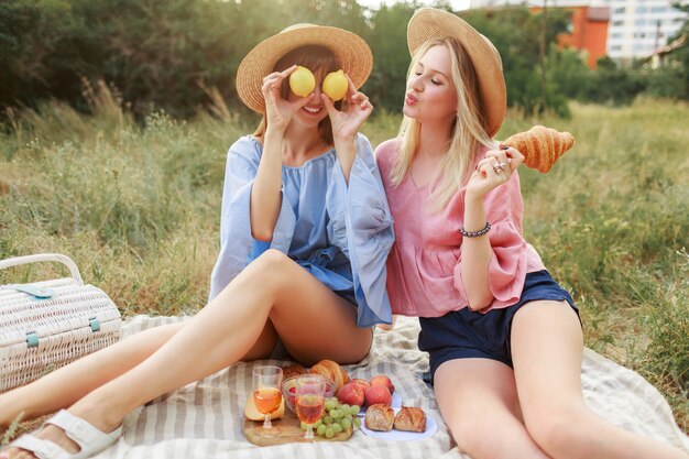 夏の公園の芝生でポーズをとって、おいしい料理、クロワッサン、ワインを楽しんでいる素敵なきれいな女性のカップル。ピクニックを楽しんでいる友人。