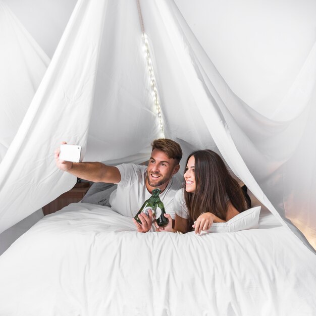 Couple lying on bed toasting alcohol bottles taking selfie on cellphone