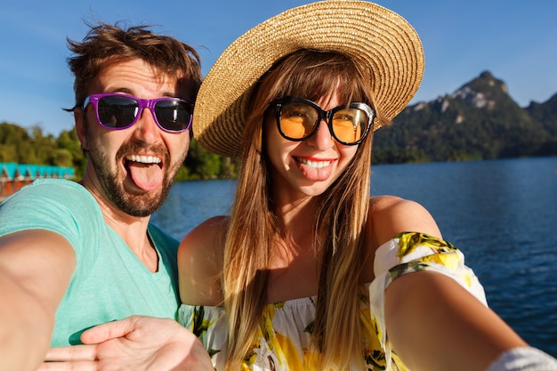 couple marking selfie near amazing lake and mountains view, wearing stylish clothes and accessories. Playful happy atmosphere.