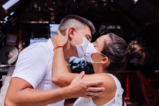 Couple in medical white face masks on vacation stand outside hotel and kissing