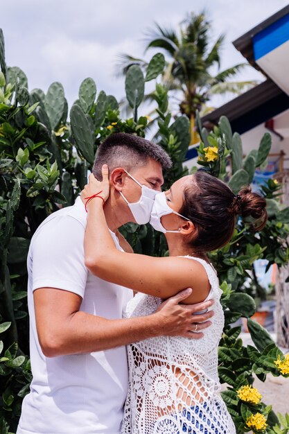 Couple in medical white face masks on vacation stand outside hotel and kissing