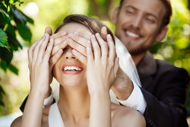 Пара молодоженов улыбается. Жених закрывает глаза невесты руками.