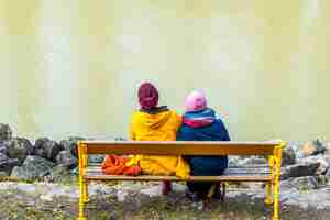 Foto gratuita coppia di persone in abiti colorati seduti su una panchina di legno gialla di fronte a un muro bianco