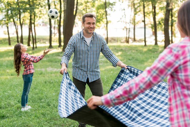 Couple placing blanket on grass near their daughter playing soccer ball in garden