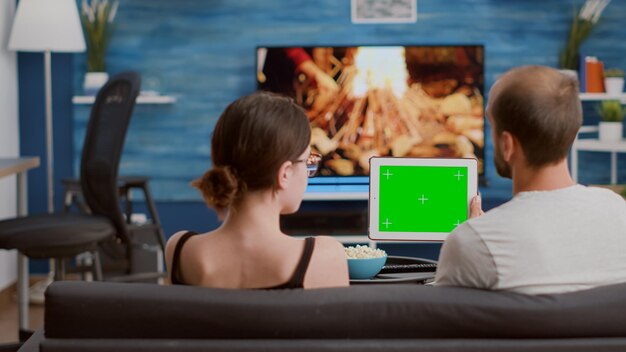 リビングルームで緑色の画面とデジタルタブレットを使用してソーシャルメディアのビデオコンテンツを見てソファでリラックスしたカップル。クロマキーデバイスでインフルエンサーvlogを見て自由な時間を楽しんでいる男性と女性。