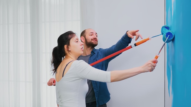 Couple repainting apartment wall with blue paint using roller brush. Apartment redecoration and home construction while renovating and improving. Repair and decorating.