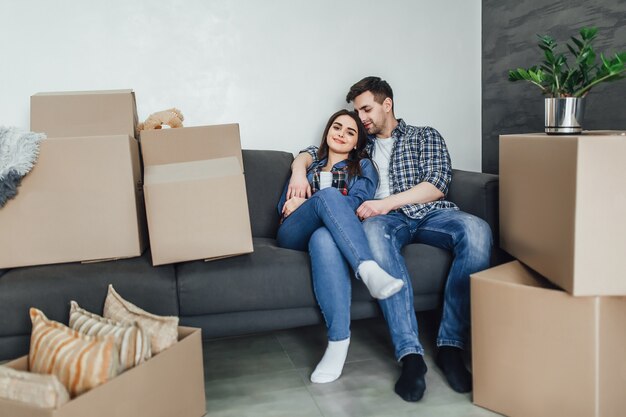 Couple resting on couch after moving in, man and woman relaxing on sofa just moved into apartment with cardboard boxes on floor