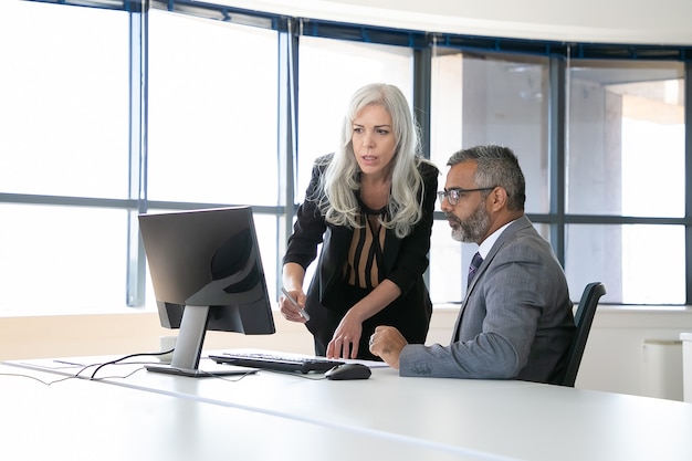 Couple of serious colleagues discussing content on computer monitor, pointing at display and talking while sitting in meeting room with panoramic window. Business communication concept