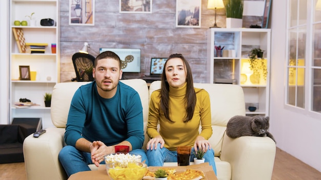 Couple sitting on the couch cheering up while watching sports on tv and eating junk food with their cat next to them.