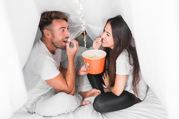 Couple sitting inside the covered bed with curtain eating popcorn