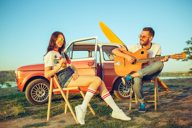 Couple sitting and resting on the beach playing guitar on a summer day near river. Caucasian man and woman