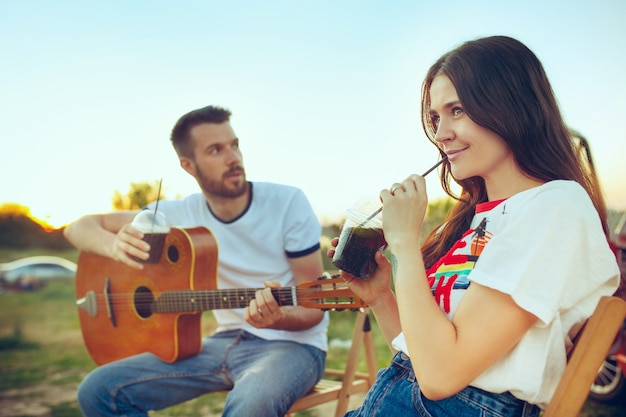 Пара сидит и отдыхает на пляже, играя на гитаре в летний день возле реки. Любовь, счастливая семья, отдых, путешествия, летняя концепция. Кавказский мужчина и женщина