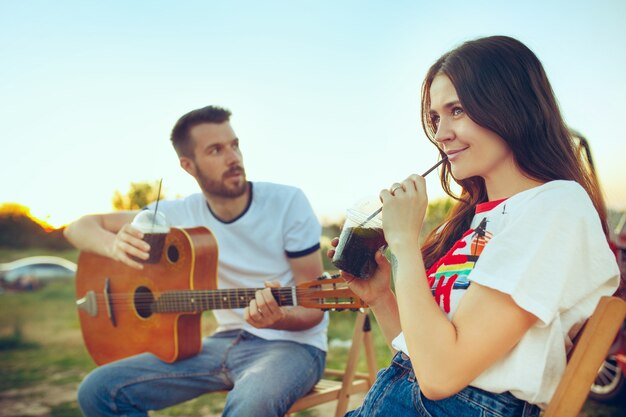 川の近くの夏の日にギターを弾いてビーチに座って休んでいるカップル。愛、幸せな家族、休暇、旅行、夏のコンセプト。白人の男性と女性