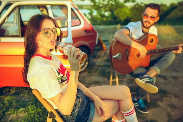 Free photo couple sitting and resting on the beach playing guitar on a summer day near river. love, happy family, vacation, travel, summer concept.