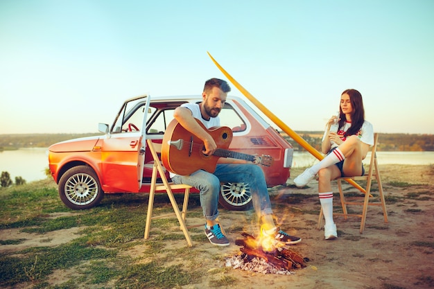 Couple sitting and resting on beach playing guitar on summer day near river