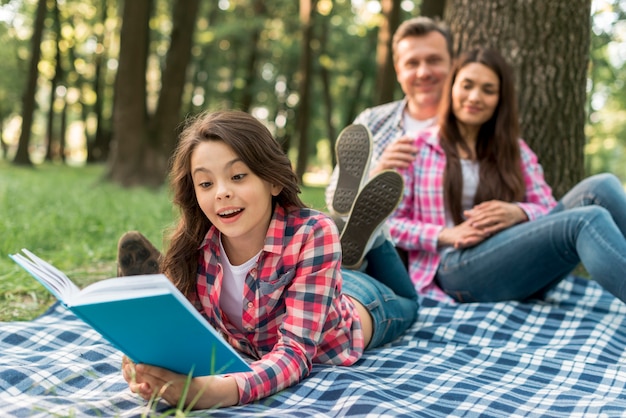 Couple sitting behind their cute girl lying on blanket reading book in park