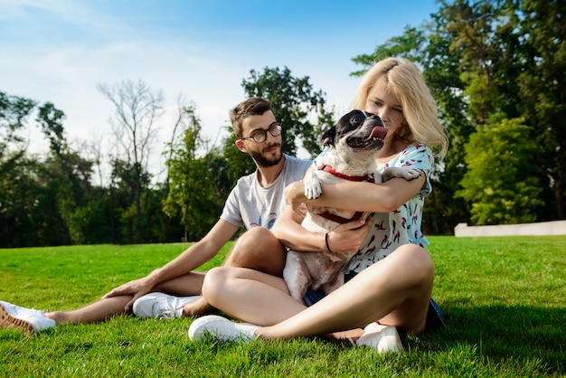 Couple smiling, sitting on grass with French bulldog in park