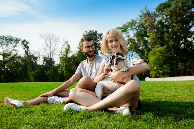 Couple smiling, sitting on grass with French bulldog in park