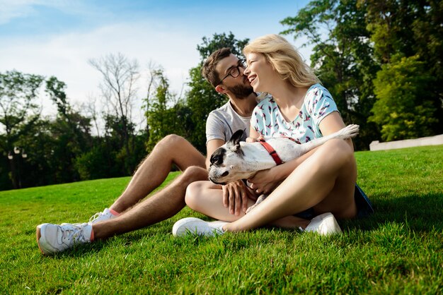 Couple smiling, sitting on grass with French bulldog in park