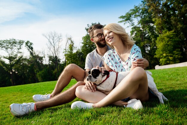 Couple smiling, sitting on grass with French bulldog in park