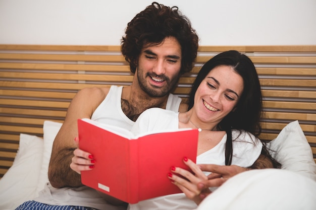 Free photo couple smiling while reading a book in bed