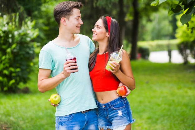 Couple standing in park holding healthy smoothies and apples looking at each other