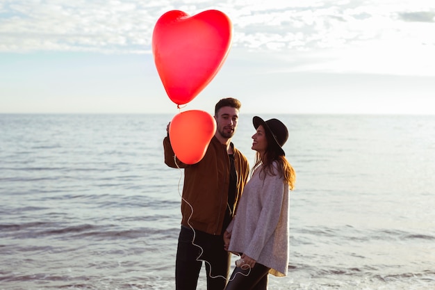 Foto gratuita coppia in piedi sulla riva del mare con palloncini cuore rosso