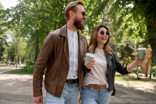 Couple in synthetic leather jackets having coffee and snack while walking outdoors