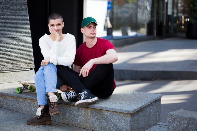 Couple in trendy clothes sitting on border near longboard