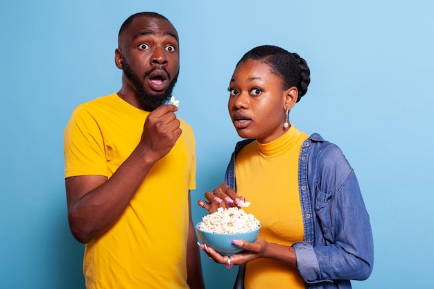 Couple watching horror movie on television and eating popcorn, being scared and terrified to watch thriller film in studio. Frightened boyfriend and girlfriend having snack and looking at tv.