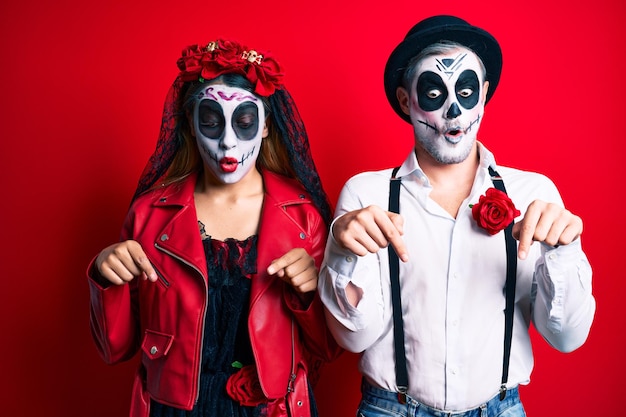 Бесплатное фото Пара в костюме дня мертвых поверх красного, указывающего вниз, с пальцами, показывающими рекламу, удивленным лицом и открытым ртом