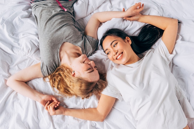 Couple of women laughing and holding hands lying in bed