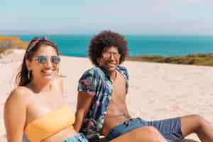 Free photo couple of young people in sunglasses sitting on beach smiling and looking at camera