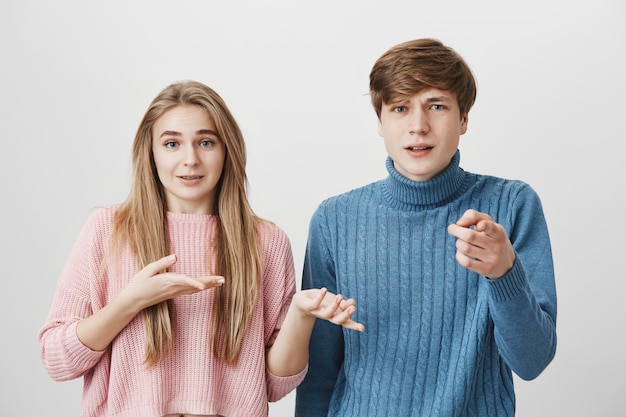 A couple of youngsters pointing with index fingers. Young fair-haired male pointing at camera with dissatisfied face expresssion