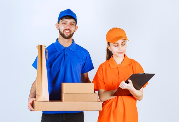 Курьерские мальчик и девочка в синей и желтой униформе держат картонные коробки для еды на вынос и пакеты для покупок и представляют список клиентов.
