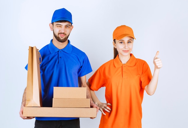 Courier boy and girl in blue and yellow uniforms holding cardboard takeaway boxes and shopping packages and showing satisfaction hand sign.