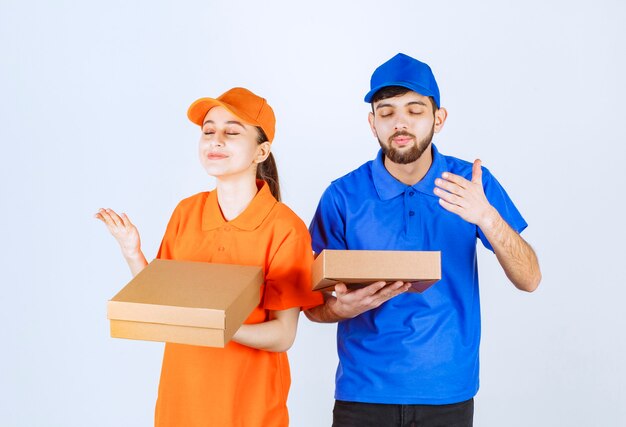 Courier boy and girl in blue and yellow uniforms holding cardboard takeaway boxes and shopping packages and smelling the food.