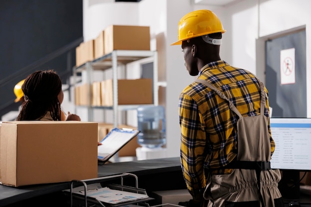 Courier checking orders list and receiving parcel in retail storehouse. African american warehouse managers preparing package delivery invoice at reception desk in storage room