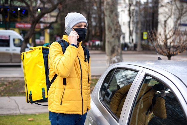 Курьер с желтым рюкзаком и черной медицинской маской возле машины разговаривает по телефону