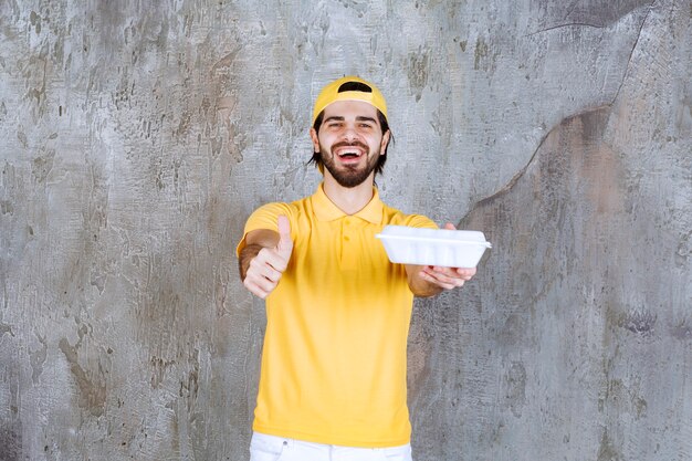 Курьер в желтой форме доставляет пластиковую коробку для еды и показывает знак рукой.